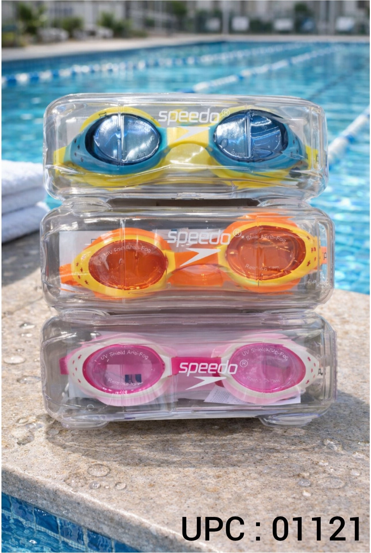 Three pairs of Speedo swim goggles stacked in clear plastic cases on a poolside ledge, with blue water and a blurred pool background.