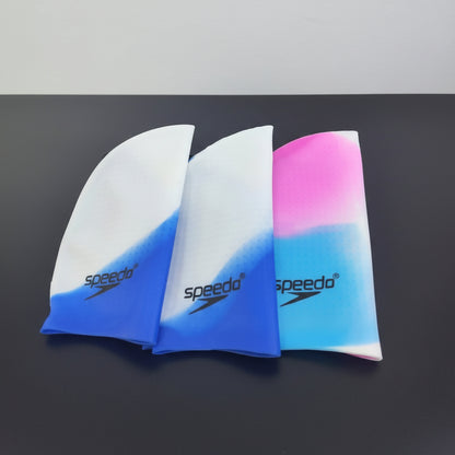 Three Speedo swim caps displayed on a dark surface, featuring white-to-blue and pink-to-blue gradient designs. A small white sign on a wooden easel reads 'WE ARE H&R', and the H&R logo appears in the top left corner.
