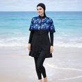 A woman wearing a black full-length swim cover-up with a blue, purple, and pink tropical leaf pattern on the upper chest and sleeves, paired with black leggings and a black head covering, standing on a beach with the ocean in the background.