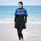 A woman wearing a black full-length swim cover-up with a blue, purple, and pink tropical leaf pattern on the upper chest and sleeves, paired with black leggings and a black head covering, standing on a beach with the ocean in the background.