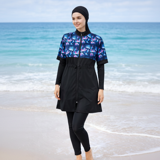 A woman wearing a black full-length swim cover-up with a blue, purple, and pink tropical leaf pattern on the upper chest and sleeves, paired with black leggings and a black head covering, standing on a beach with the ocean in the background.