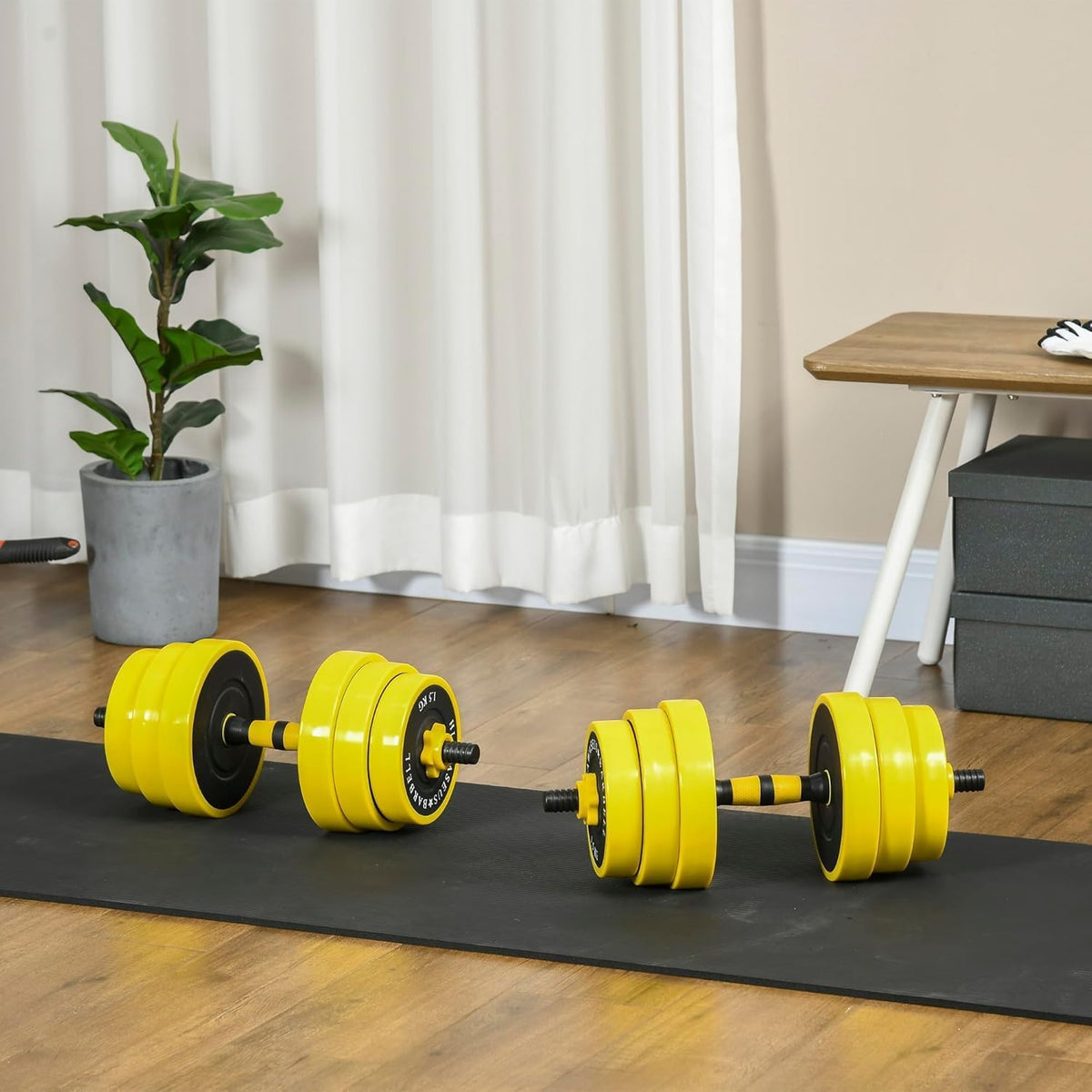 Yellow weightlifting dumbbells on a black mat with a plant and table in the background.