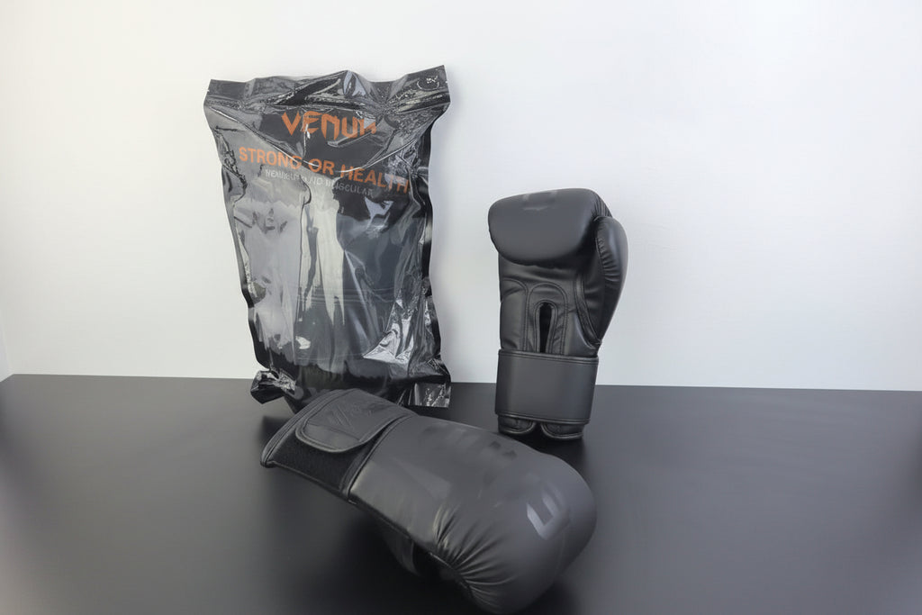 Three Venum boxing gloves in dark gray and black are displayed on a dark tabletop against a white wall. One glove is shown in its clear plastic packaging with orange Venum branding, one is standing upright, and one is lying on its side.