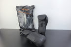 Three Venum boxing gloves in dark gray and black are displayed on a dark tabletop against a white wall. One glove is shown in its clear plastic packaging with orange Venum branding, one is standing upright, and one is lying on its side.