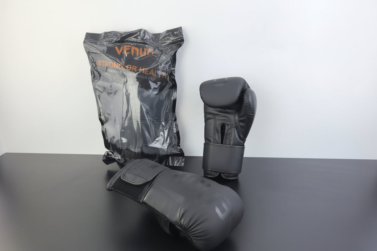 Three Venum boxing gloves in dark gray and black are displayed on a dark tabletop against a white wall. One glove is shown in its clear plastic packaging with orange Venum branding, one is standing upright, and one is lying on its side.